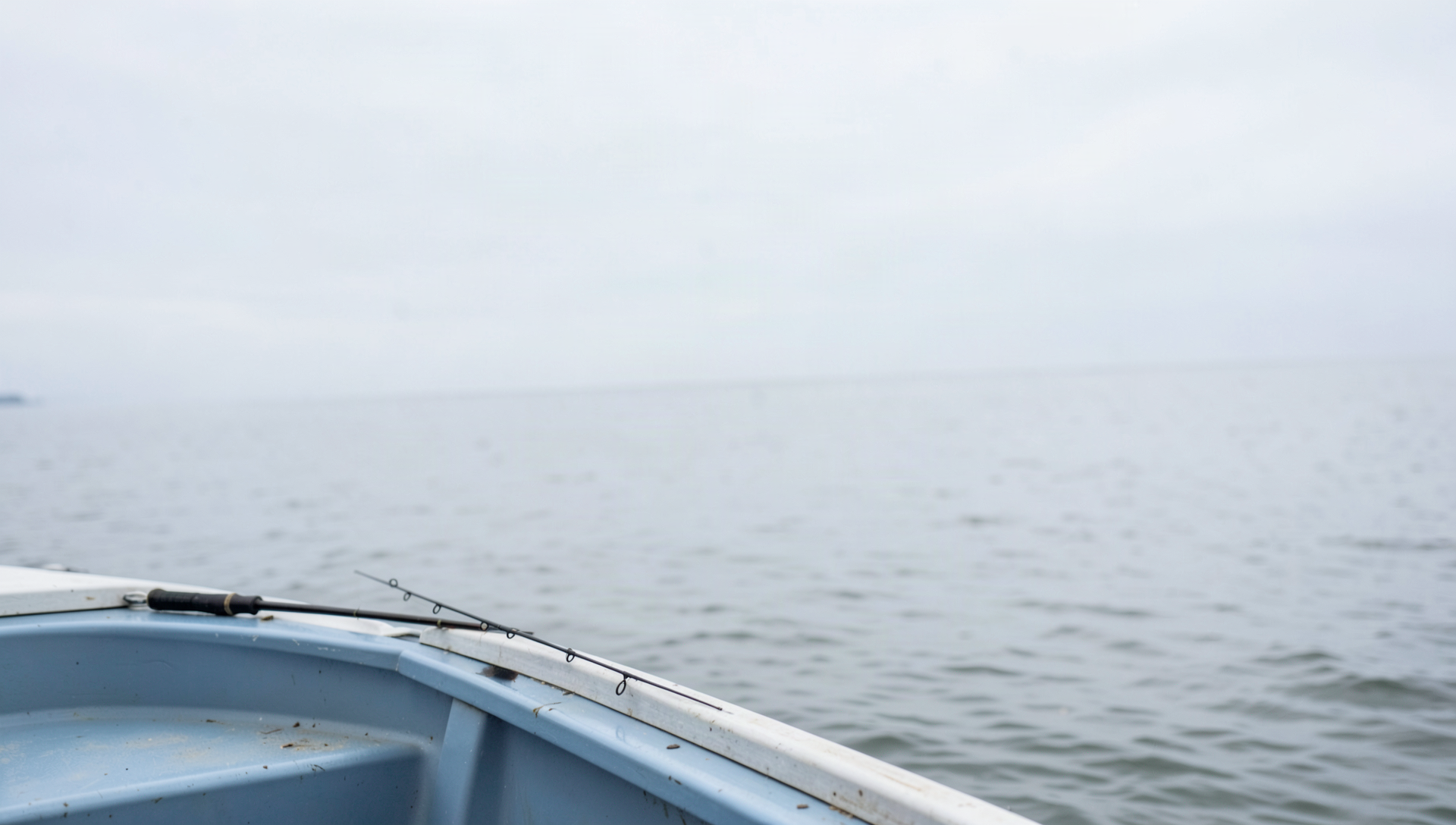 A short parabolic jigging rod lying across the gunwale of a drifting boat with calm open water and a pale overcast sky behind it